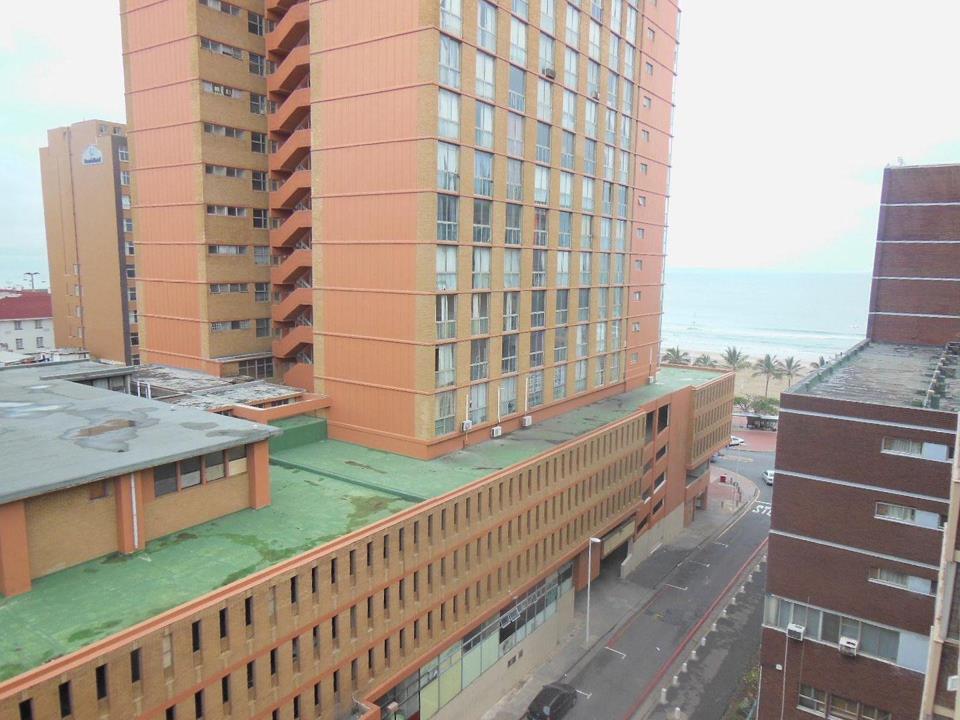 Elevated view from the apartment looking towards Durban's beachfront, showing multi-storey orange-brick buildings, a flat green rooftop, and a partial view of the Indian Ocean and sandy beach in the background.