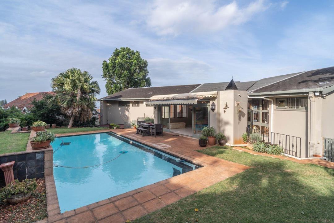Rear garden view of a double-storey Upper Glenwood home showing a rectangular swimming pool with brick coping, a covered patio with outdoor dining set, mature palm and shade trees, and the house façade with an awning and