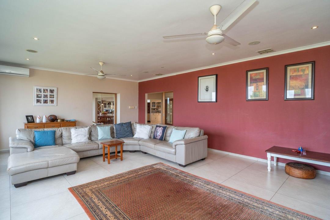 Spacious ground-floor lounge with large L-shaped cream leather sectional sofa, Persian-style area rug, ceiling fan, recessed downlights, air-conditioning unit, feature terracotta-red accent wall with framed artworks, and