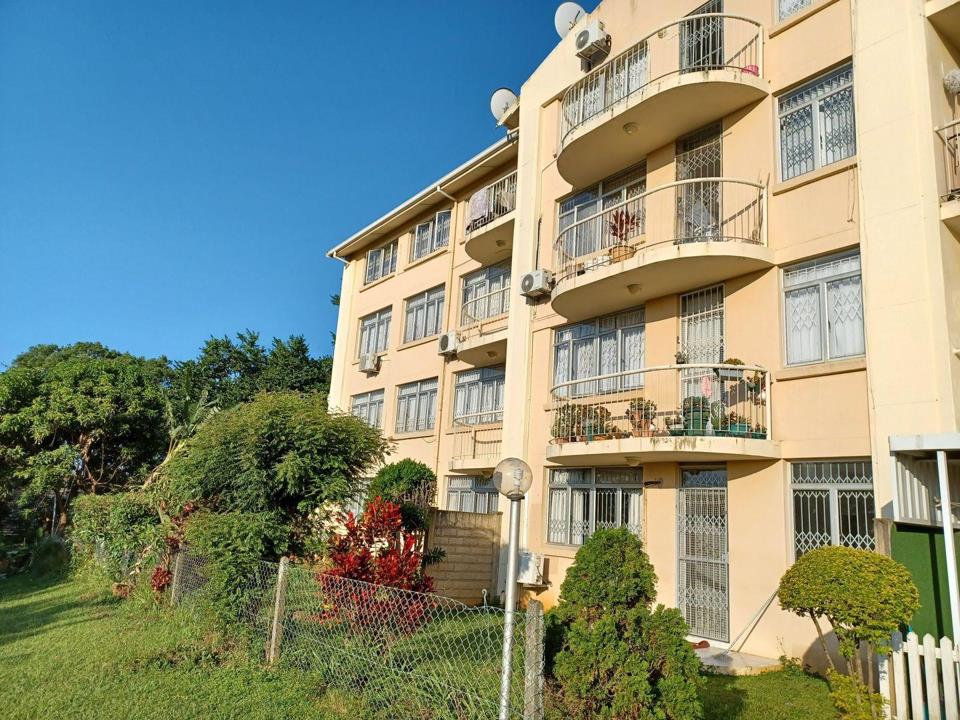 Exterior view of a three-storey cream-rendered apartment block in Sherwood, Durban, showing curved balconies, security gates, air-conditioning units, and a manicured garden with tropical shrubs under a clear blue sky.