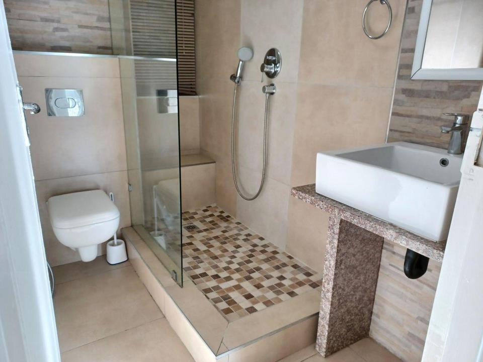 Renovated bathroom featuring a wall-mounted WC, frameless glass shower enclosure with mosaic-tile floor pan, vessel basin on a granite-faced vanity counter, and warm large-format neutral wall tiles with a slate-look feat