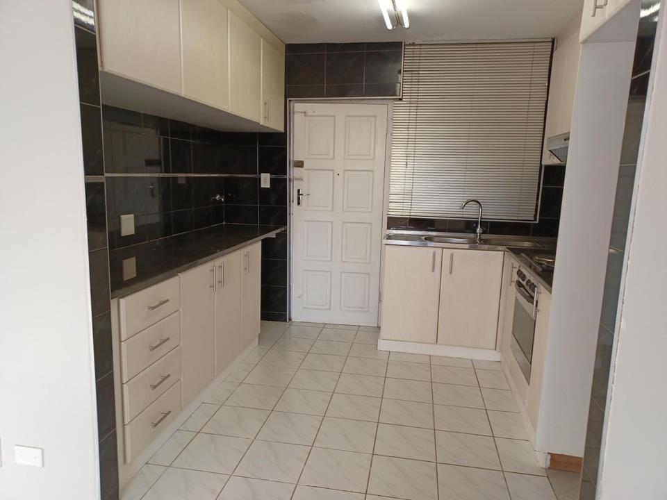 Galley-style kitchen with cream flat-panel cabinetry, dark granite-effect countertops, glossy black tile splashback, stainless-steel sink, freestanding stove, and white floor tiles under fluorescent strip lighting.