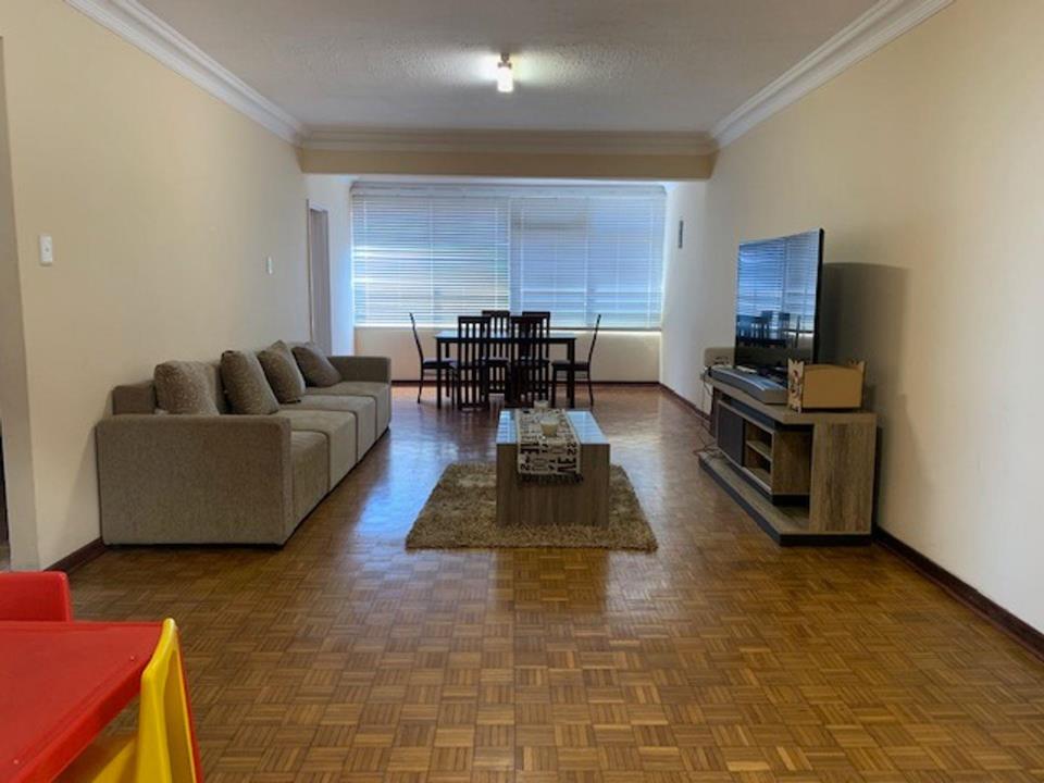 Spacious open-plan lounge and dining area with parquet block flooring, large grey L-shaped sofa, wooden coffee table on a shag rug, flat-screen TV on a media unit, dining table and chairs in the background, and large Ven