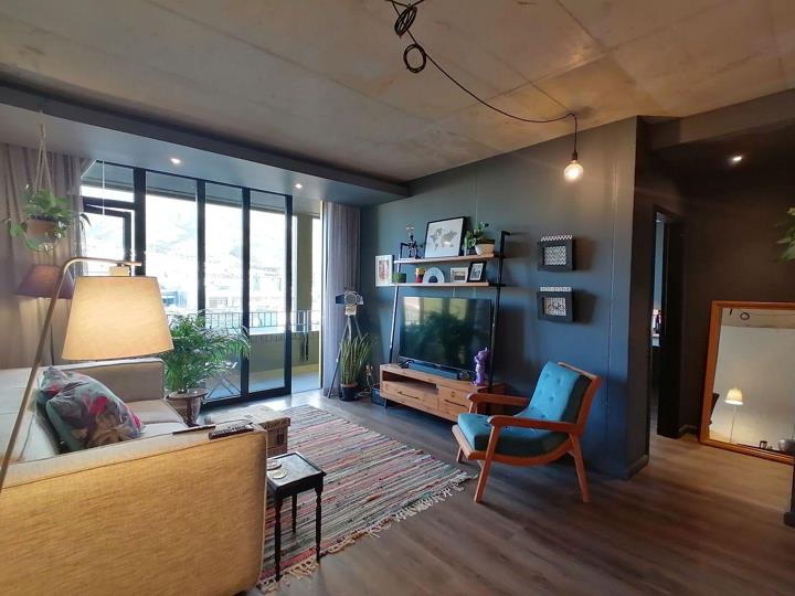 Lounge area viewed toward the balcony, featuring a deep teal accent wall with a lean-to shelving unit and large flat-screen TV on a wooden media unit, a mid-century teal button-back armchair, a beige sofa with scatter cu