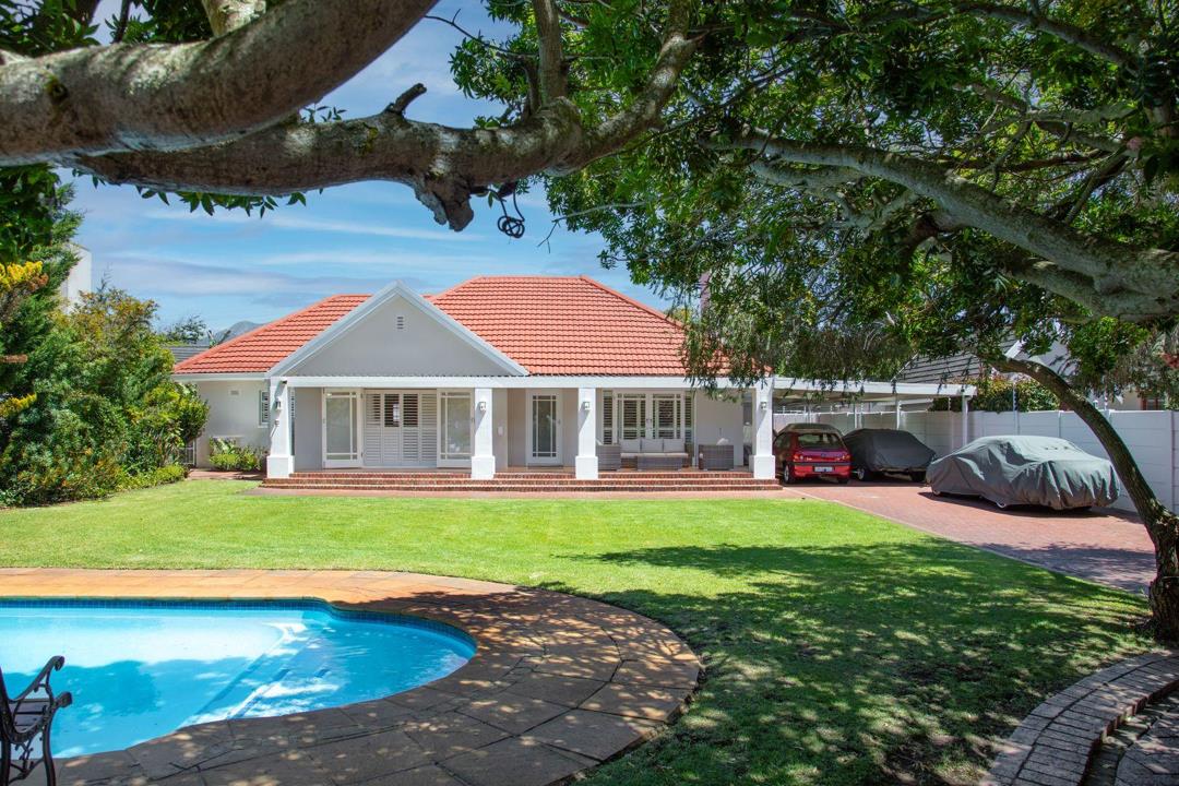 Front exterior of a single-storey white house with a terracotta tile roof, wide covered verandah, manicured lawn, a freeform swimming pool in the foreground, and two cars parked under a carport to the right, photographed
