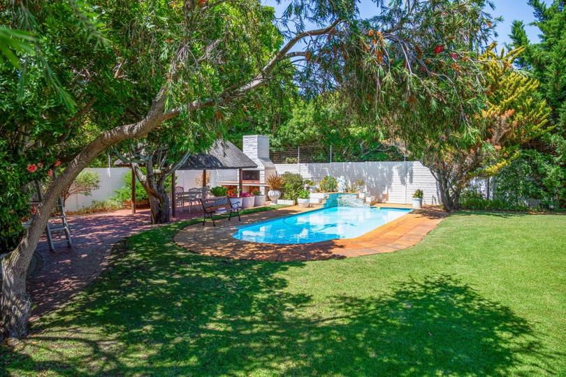 Rear garden view showing a rectangular swimming pool with a tiled surround, a covered lapa with a built-in braai and outdoor dining furniture, and lush mature trees enclosing the space, with a well-maintained lawn in the