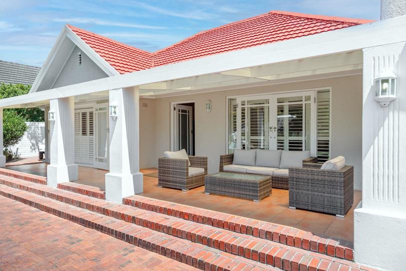 Covered front verandah with white structural columns, a terracotta tile roof, rattan sofa and armchairs on a tiled floor, louvred plantation shutters on the façade, and brick steps leading down to the paved driveway.