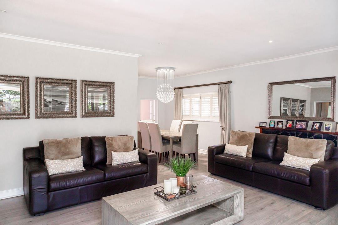 Open-plan lounge and dining room with laminate flooring, two dark-brown leather sofas, a grey wooden coffee table, a crystal pendant chandelier above a six-seater dining table, ornate wall-mounted mirrors, and plantation