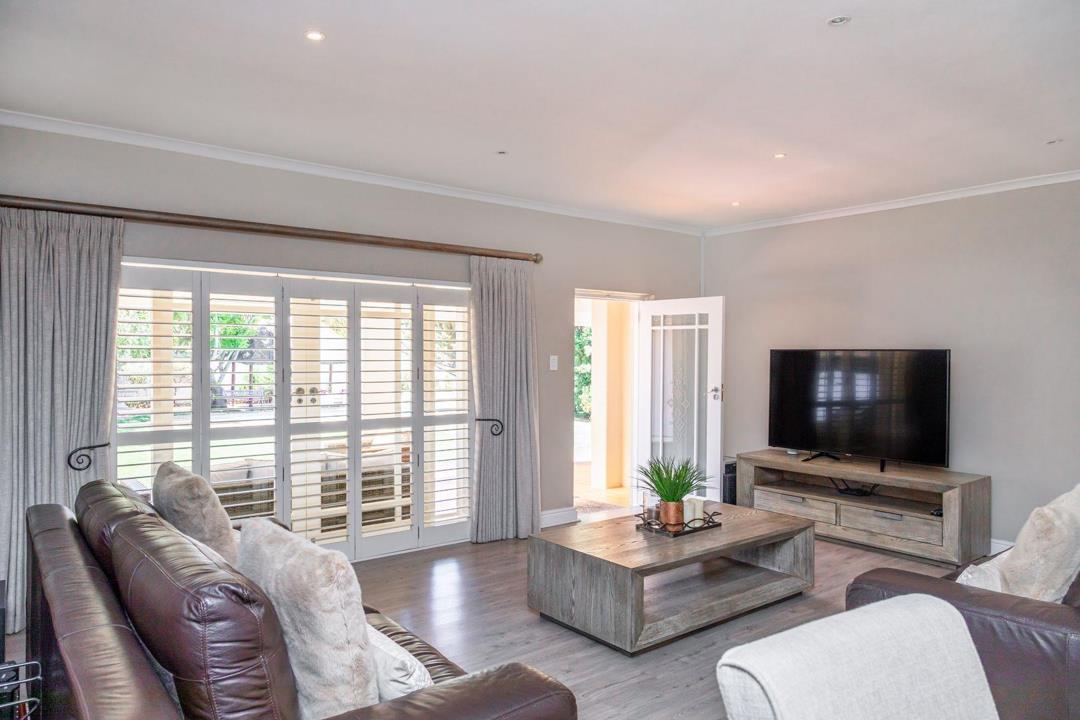 Lounge with laminate flooring, dark-brown leather sofas, a flat-screen television on a wooden media unit, louvred plantation shutters and full-length curtains on a set of French doors opening to the verandah and garden b