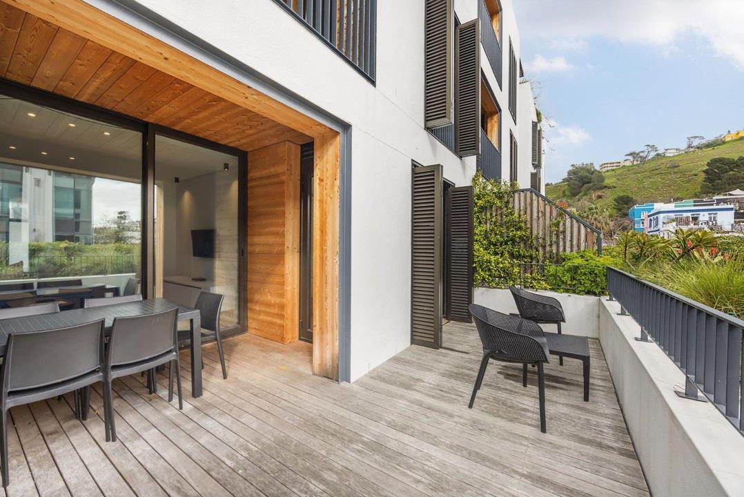 Spacious timber-decked balcony of Unit 201 at Old Cape Quarter showing an outdoor dining set on the left, two lounge chairs to the right, cedar-lined sliding door entrance to the living area, dark louvred shutters on the