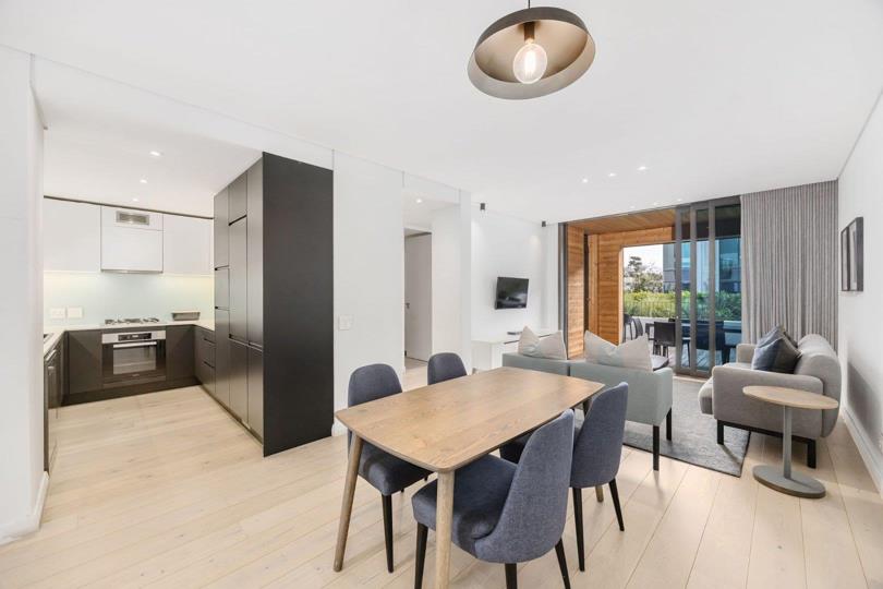 Open-plan kitchen, dining, and living area with pale OGGIE wood flooring, a wooden dining table with six blue upholstered chairs under a dome pendant light, dark matte kitchen cabinetry, built-in oven, and sliding glass 