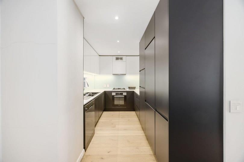 Close-up view down the U-shaped kitchen corridor showing dark matte flat-panel lower cabinets, white upper cabinets, glass splashback, stainless-steel gas hob, built-in oven, integrated dishwasher drawer, and pale engine