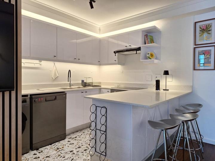White gloss U-shaped kitchen with LED cove lighting in a stepped ceiling recess, stone-look island counter with two bar stools, built-in wire wine rack beneath the island, stainless steel dishwasher, extractor hood, open