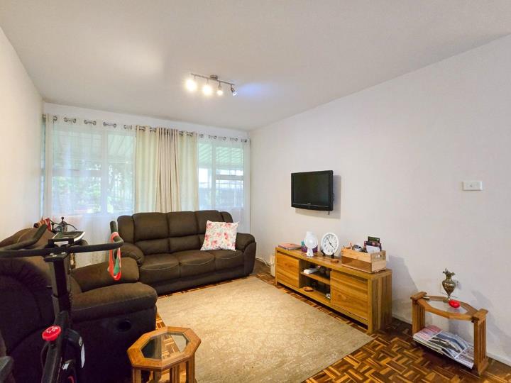 Living room with warm parquet flooring, a brown recliner sofa suite, wall-mounted flat-screen TV on a timber media unit, sheer curtained window, and track lighting on a white ceiling.