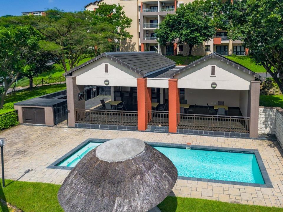 Eye-level view of the complex swimming pool with a thatched umbrella in the foreground and a large covered lapa featuring outdoor dining furniture, red-painted columns, and dark tile flooring behind.