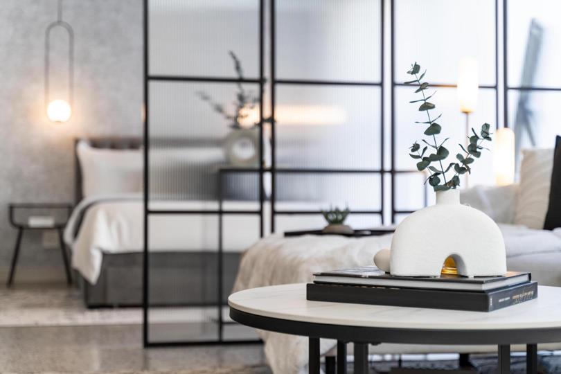Close-up detail of a white marble-top coffee table styled with a ceramic dome vase holding eucalyptus stems, with the steel-framed ribbed-glass bedroom partition and a double bed dressed in white linen softly visible in 