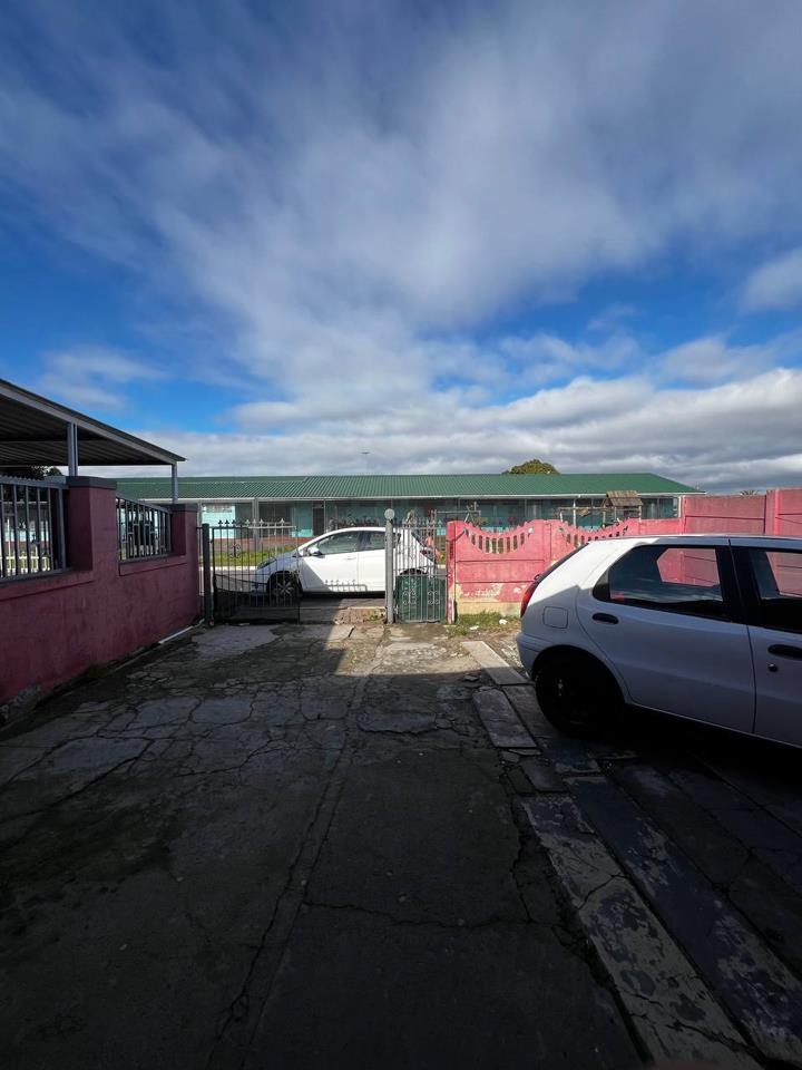 Rear yard perspective looking toward the street gate, flanked by pink boundary walls on both sides, with two white vehicles parked on cracked concrete paving and a neighbouring single-storey building with a green metal r