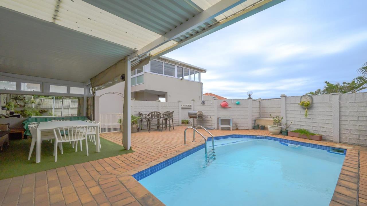 Covered patio with white outdoor dining table and chairs on an artificial-grass mat adjoins a blue-tiled rectangular swimming pool surrounded by brick paving, with a second storey glazed sunroom visible in the background