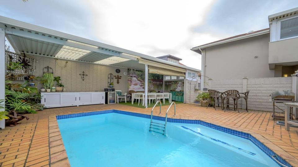 View across the swimming pool toward the covered entertainment patio, showing a white outdoor sideboard with a bar fridge, hanging plants, decorative wall cross, cast-iron bistro chairs, and a braai station to the right.