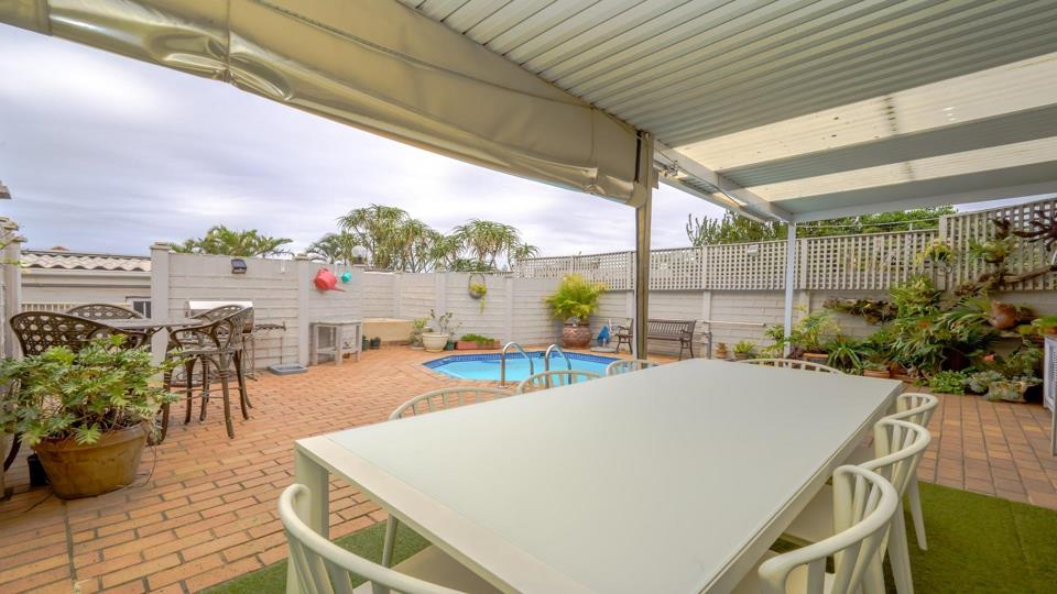 White outdoor dining table and chairs under a retractable awning on a brick-paved courtyard, looking toward the swimming pool with lattice boundary walls, palm trees, and established garden planting.