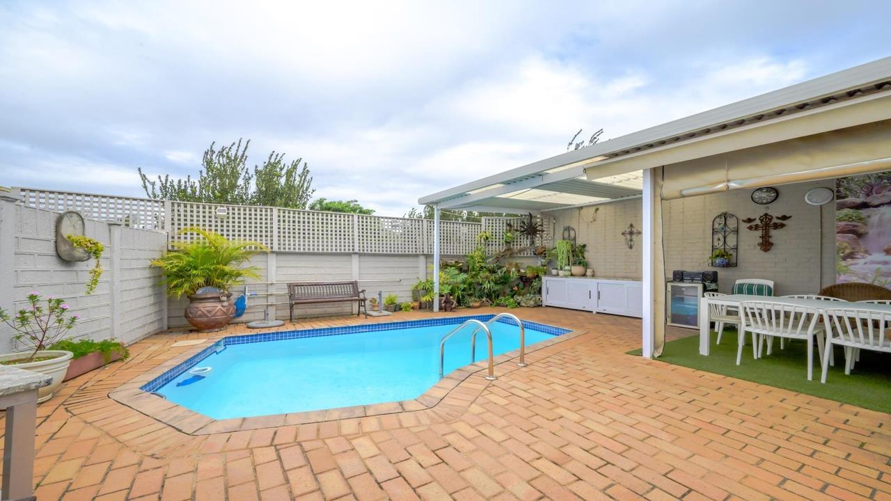 Full view of the shared swimming pool courtyard with brick paving, garden bench, large terracotta pots with tropical plants, and the covered entertainment area to the right featuring white outdoor seating and decorative 