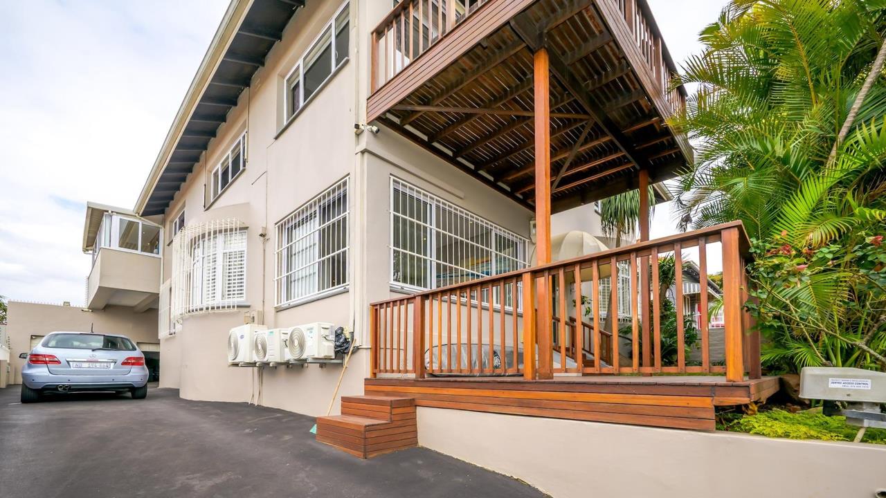 Exterior side elevation of a double-storey rendered house showing a timber deck with wooden balustrade and steps, burglar-bar-fitted windows, two external air-conditioning units, a parked sedan, and lush tropical garden 
