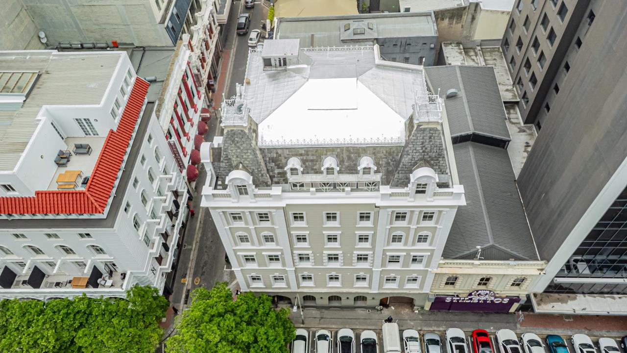 Aerial drone view directly above a five-storey heritage building in Cape Town City Centre, showing a freshly painted grey-and-white mansard roof with arched dormers, ornamental ironwork balustrades, and a flat white uppe