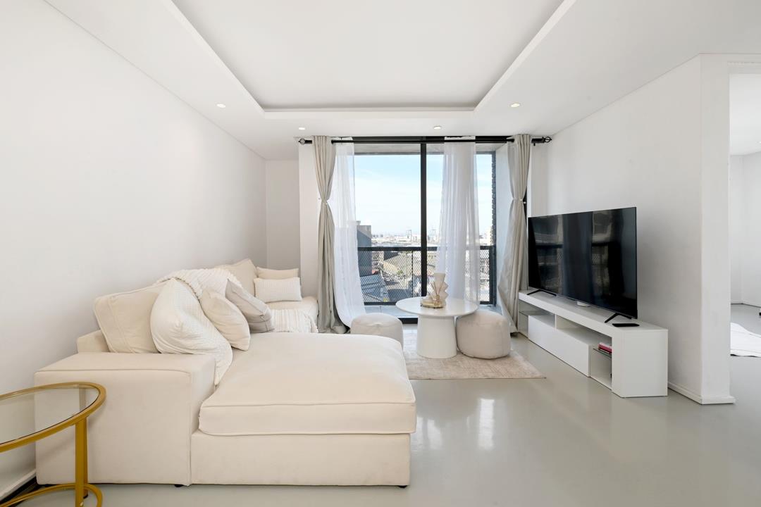 Bright open-plan lounge with oversized cream sectional sofa, polished grey floors, coffered white ceiling with downlights, wall-mounted flat-screen TV on white media unit, and floor-to-ceiling sliding doors opening to a 