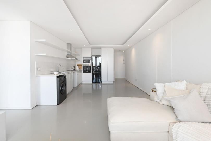 Wide-angle view of the open-plan kitchen and living area showing the full depth of the apartment, with white flat-panel kitchen units on the left, polished concrete-effect floors throughout, and a cream lounge suite visi