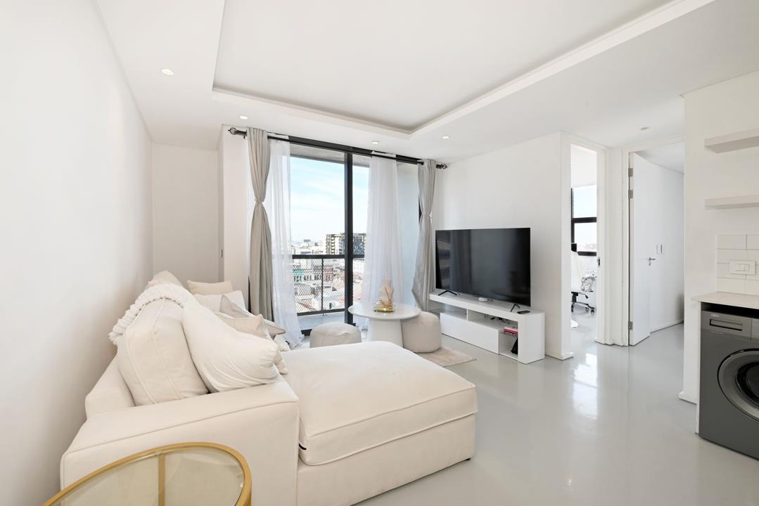 Living room viewed from behind the sofa showing the balcony sliding doors with sheer curtains framing a city skyline view, wall-mounted TV, white media unit, and a doorway leading to a study or second bedroom with an off