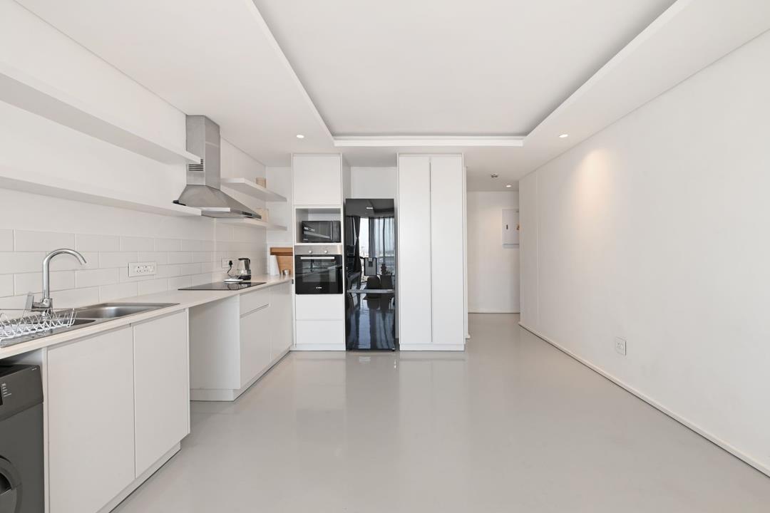 Kitchen viewed from the sink end showing the stainless-steel extractor, built-in oven and microwave column, tall white integrated refrigerator housing, and a long open corridor leading toward the balcony and living area 