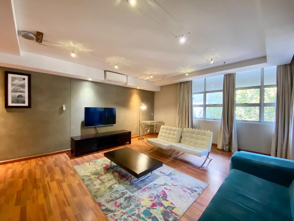 Open-plan lounge with warm timber parquet flooring, coffered ceiling with track lighting, wall-mounted flat-screen TV on a feature grey wall, a teal velvet sofa, two white Barcelona-style chairs, a black coffee table on 
