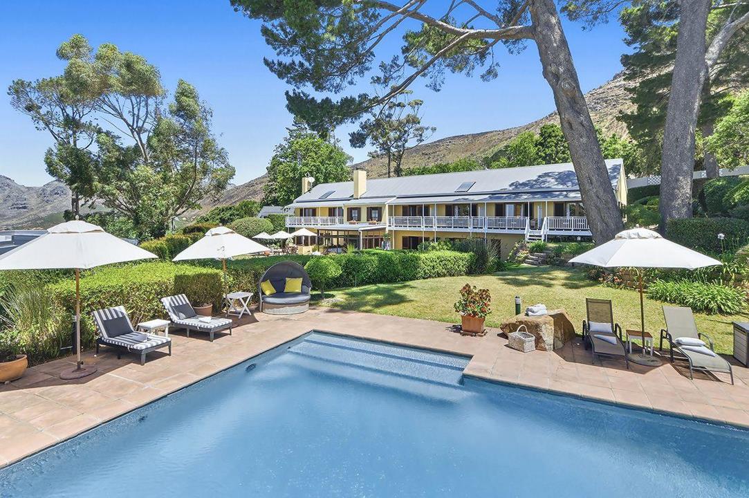 Aerial-perspective rear view of a double-storey yellow Cape colonial house with a large rectangular swimming pool, tiled sun terrace with sun loungers and parasols, manicured lawn, and a dramatic mountain backdrop under 