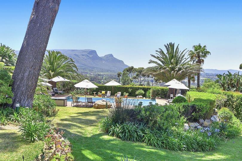 Wide garden view looking toward the swimming pool and terrace from a landscaped lower garden, with tall pine trees, palm trees, mixed garden beds, a jacuzzi unit to the left, and a flat-topped mountain range visible on t