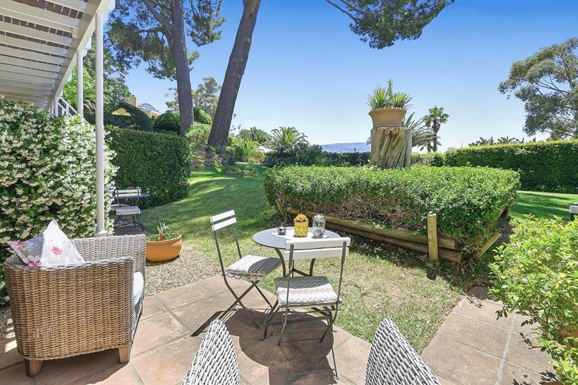 Covered ground-floor patio with wicker armchairs and a small bistro table set for two, overlooking a neat lawn bordered by jasmine and clipped hedging, with tall pines and a glimpse of ocean in the distance.