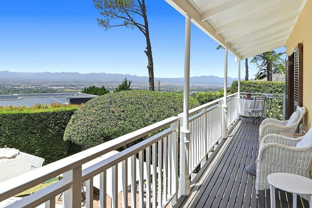 Upper-storey wraparound verandah with white timber balustrade and columns, furnished with white wicker chairs and a bistro table, overlooking a sweeping panorama of the Cape Flats, mountain ranges and distant valley belo