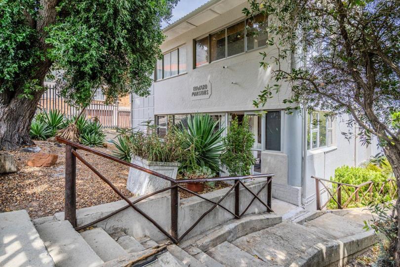 Front exterior of Edward Mansions, a two-storey white rendered apartment block with large windows, surrounded by mature trees and indigenous plantings, with a concrete staircase and metal handrail leading to the entrance