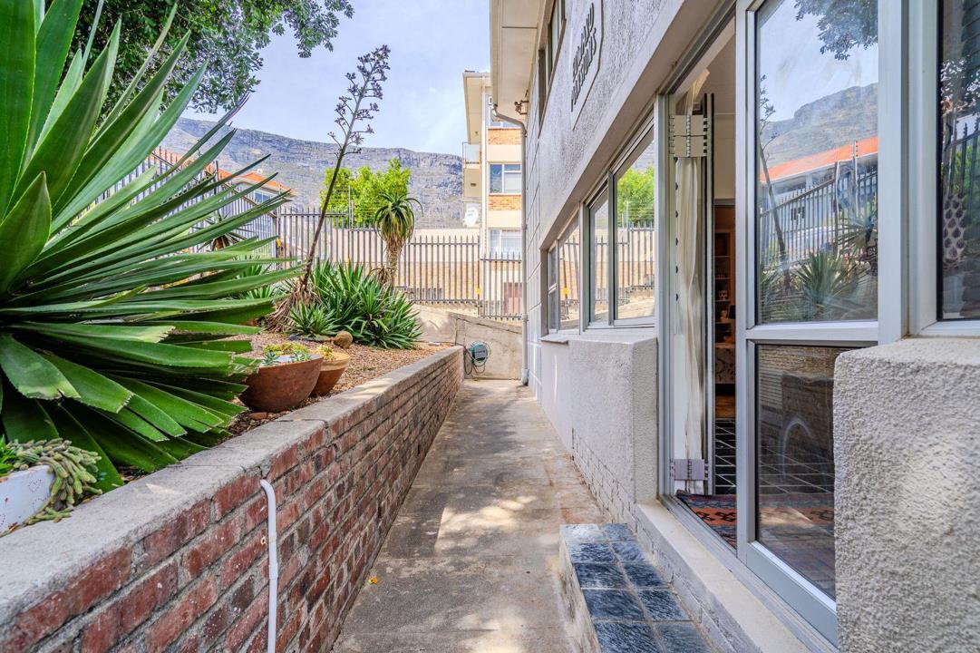 Side passage of Edward Mansions showing a raised brick planter bed with aloes and succulents, a concrete walkway alongside the rendered white building facade, and Table Mountain visible in the background.