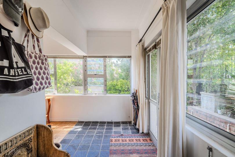 Glass-enclosed entrance sunroom with mixed slate-tile and timber parquet flooring, sheer white curtains, multiple aluminium-framed windows overlooking a green garden, and coat hooks with bags on the left wall.