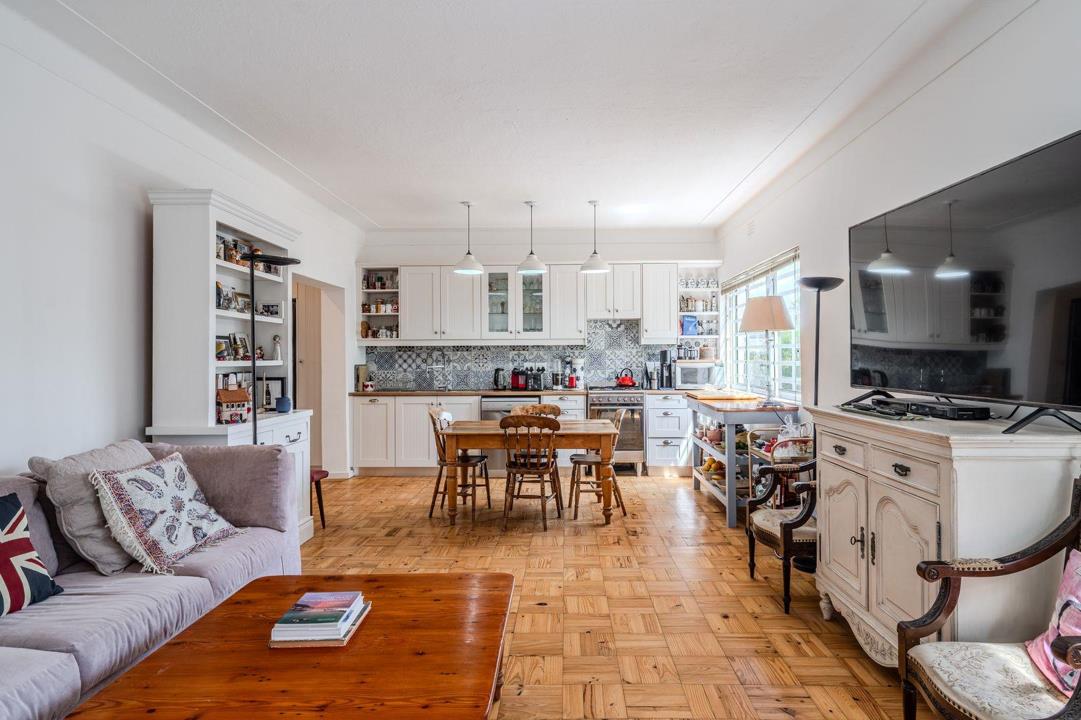 Spacious open-plan living, dining and kitchen area with parquet timber flooring, white shaker kitchen cabinetry with patchwork tile splashback, pendant lighting, a rustic wooden dining table, and a large flat-screen TV i