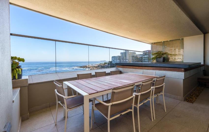 Covered balcony dining area with a six-seat timber-topped outdoor table and white frame chairs, built-in stone-faced outdoor kitchen counter, glass balustrade, and a panoramic Atlantic Ocean and coastline view.