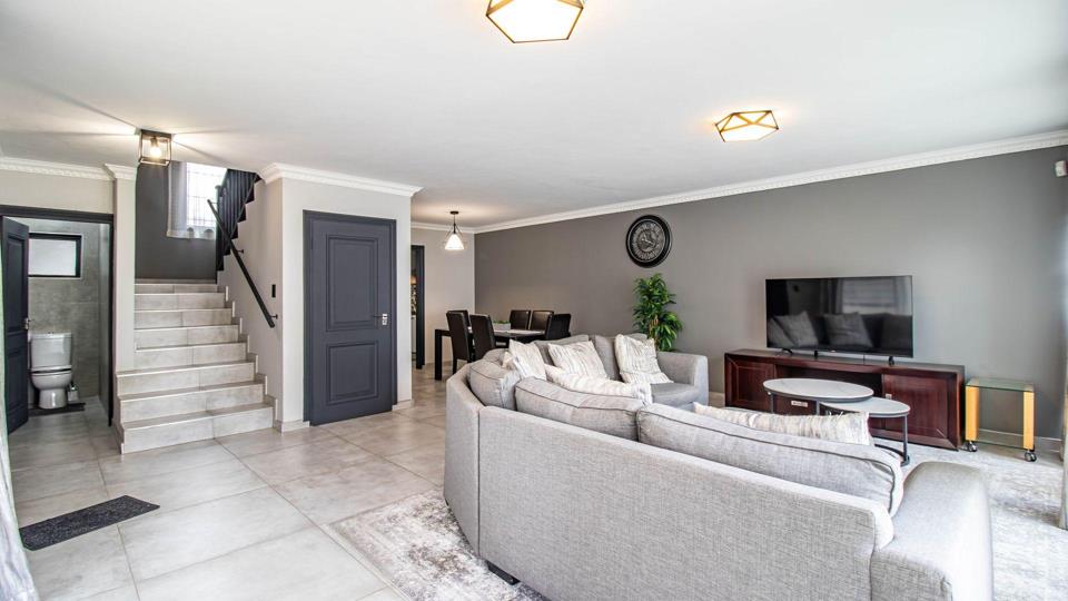 Open-plan ground-floor living area featuring a large grey corner sofa, flat-screen TV on a timber unit, grey accent walls with white cornicing, geometric gold pendant lights, large-format porcelain tiles, and a staircase