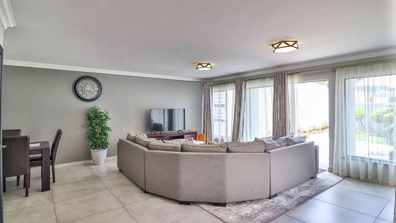 Living and dining area showing a grey corner sectional sofa, flat-screen TV, wall clock, potted plant, and a dining table with dark chairs to the left, with a sliding glass door opening to the garden and grey walls with 
