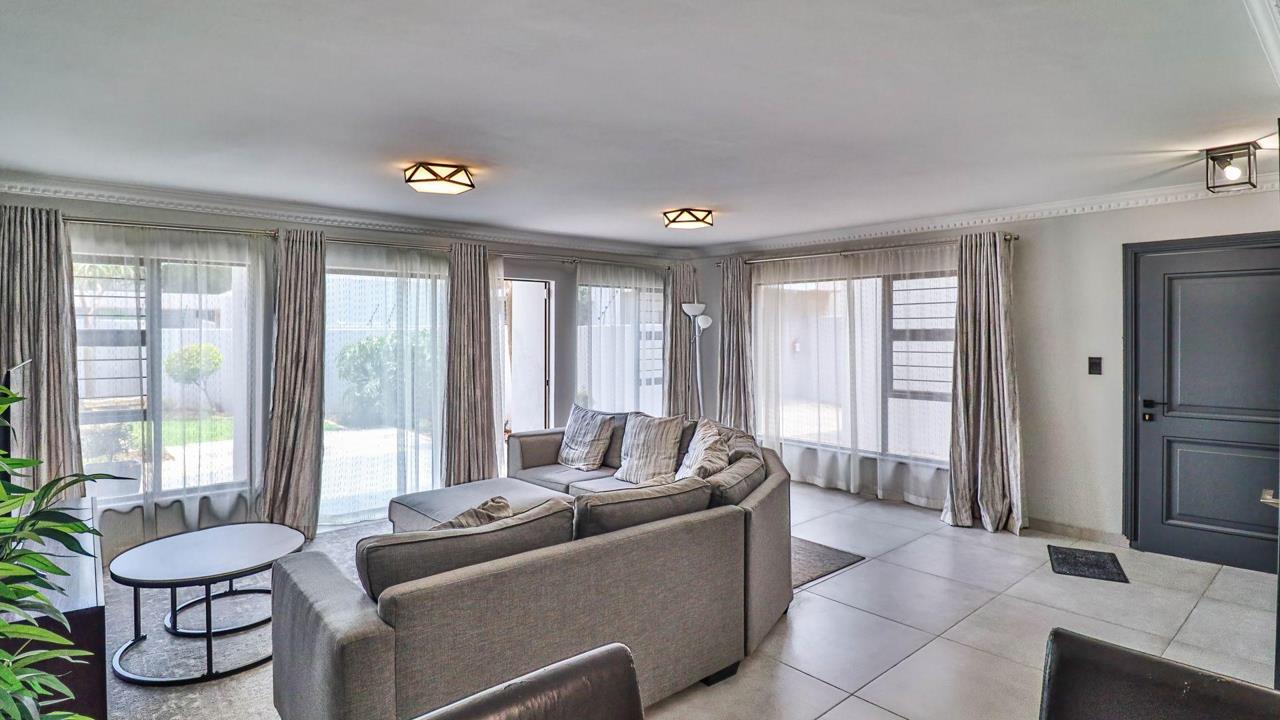 Spacious lounge viewed from the garden side, with three sets of large aluminium-framed sliding doors and windows dressed in sheer grey curtains flooding the room with natural light; a grey corner sectional sofa faces a s