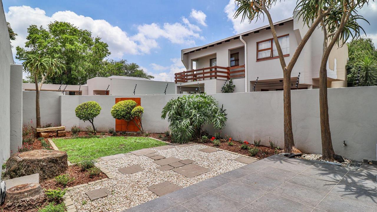 Rear garden view of a double-storey duplex unit in Melville, showing a paved courtyard with slate stepping stones set in white pebbles, manicured lawn, shaped topiary, established trees, and plastered boundary walls unde