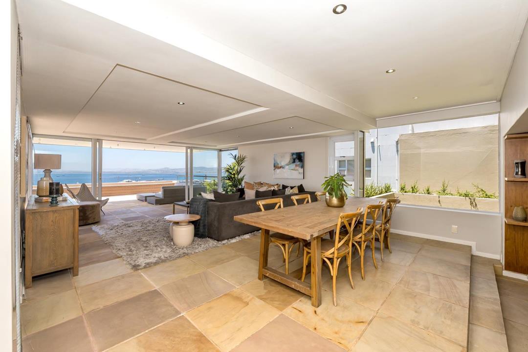 Open-plan dining and living area featuring a large reclaimed-wood dining table with cross-back chairs for eight, a shag-rug lounge zone with grey corner sofa, coffered ceiling, and expansive ocean views through floor-to-