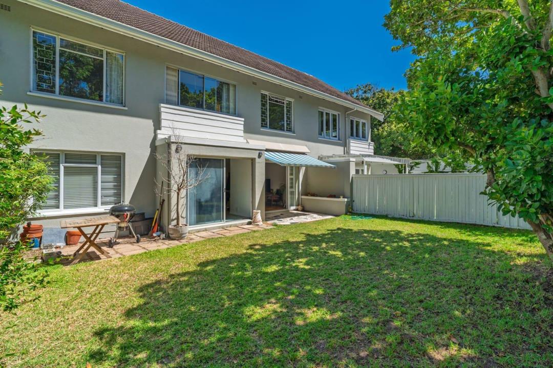 Rear exterior of a double-storey plastered apartment complex showing a well-maintained lawn garden, paved patio with Weber braai, retractable blue-and-white striped awning over sliding glass doors, and a secure white tim