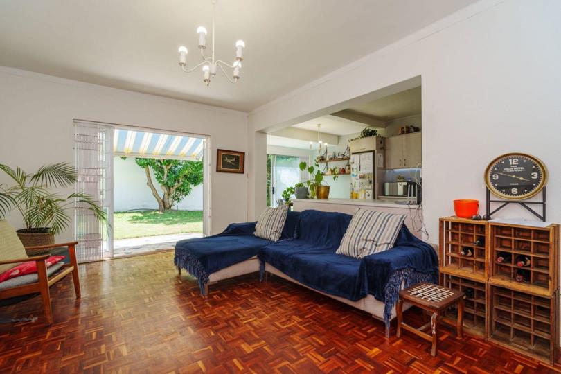 Open-plan lounge with rich parquet timber flooring, a blue sectional sofa, chandelier ceiling light, and sliding glass doors opening to a private patio and garden; an open-plan kitchen with stainless-steel fridge is visi