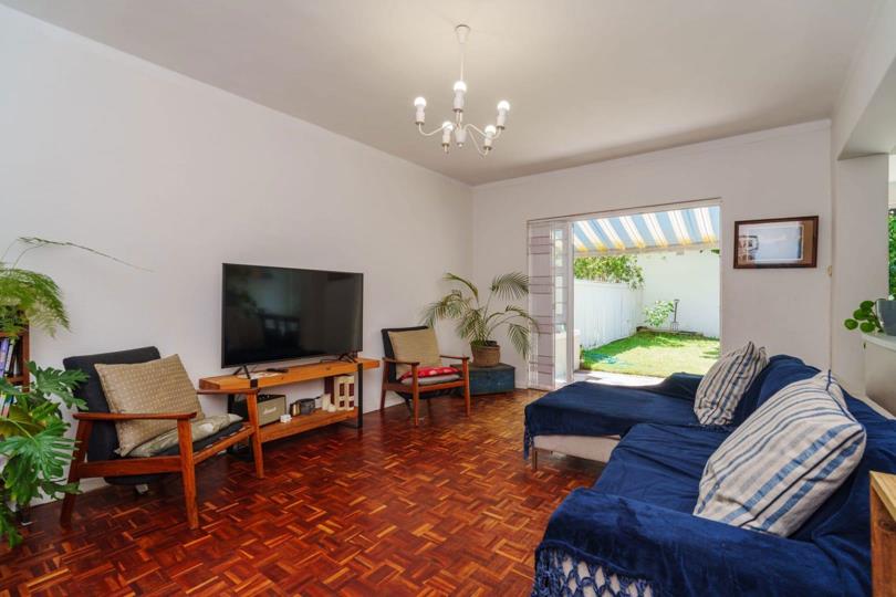 Lounge area viewed from the opposite angle, showing a large flat-screen television on a timber stand, mid-century timber armchairs, blue sectional sofa, warm parquet floors, chandelier, and sliding glass doors leading to