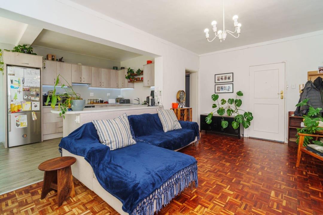 Open-plan lounge and kitchen showing light wood flat-panel cabinetry with under-counter LED strip lighting, a stainless-steel French-door fridge, breakfast counter overhang, parquet timber flooring throughout, and a chan